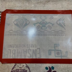 Op de afbeelding: Een rechthoekige rode siliconen bakmat met een wit gaasoppervlak. De mat heeft een rode rand en is ontworpen voor gebruik in de oven. De mat ligt op een getextureerd oppervlak.