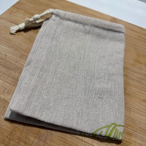 May include: A small, beige drawstring bag made from textured fabric. The bag features a green mountain-like graphic on the bottom right corner. The drawstring is a natural, off-white colour.