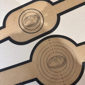 Op de afbeelding: Twee bruine siliconen bakmatten met zwarte randen. Eén mat heeft een cirkel met concentrische cirkels en metingen in inches en centimeters. De andere mat heeft een enkele cirkel met een illustratie van een brood. Beide matten hebben een illustratie van een brood.