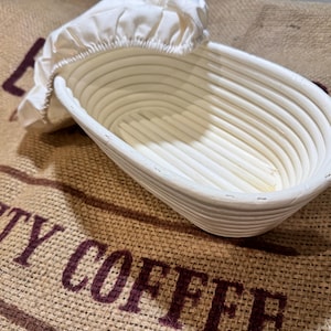 May include: Oval white wicker bread proving basket with a white linen liner. The basket is empty and rests on a burlap sack printed with the words "SPECIALTY COFFEE" in dark red.
