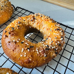 Może przedstawiać: Świeżo upieczony, złotobrązowy bagel posypany sezamem, makiem i czarnymi ziarnami sezamu. Bagel leży na czarnym drucianym ruszcie.