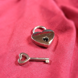 May include: A silver heart-shaped padlock with a keyhole and a matching silver key. The padlock and key are on a red fabric background.
