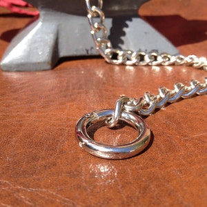 Op de afbeelding: Een zilveren ketting met een ronde, zilveren ring eraan. De ketting ligt op een bruin lederen oppervlak.