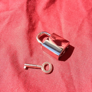 Puede incluir: Un candado plateado y una llave sobre un fondo de tela roja. El candado tiene un acabado cromado brillante y un diseño sencillo. La llave es pequeña y tiene una forma tradicional.