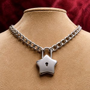 May include: A silver-toned metal chain necklace featuring a polished star-shaped padlock pendant. The padlock has a smooth, reflective surface with a central keyhole cutout. The thick, interlocking chain links connect directly to the padlock shackle.