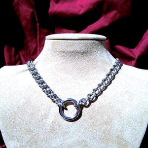 Op de afbeelding: Een zilverkleurige ketting met dikke, in elkaar grijpende ovale schakels. De ketting wordt gecentreerd door een grote, ronde metalen ringsluiting die de twee zijden van de ketting met elkaar verbindt voor een opvallende, uniforme uitstraling.