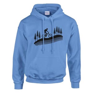Puede incluir: Una sudadera con capucha azul claro con una silueta negra de una persona montando una bicicleta de montaña en un camino sinuoso a través de un bosque. El texto "Gildan" es visible en el interior de la capucha.