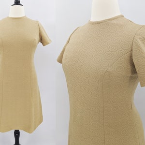 May include: A tan-colored, short-sleeved dress with a round neckline. The dress features a textured pattern of concentric circles across the fabric. The dress is displayed on a mannequin, showcasing its A-line silhouette and tailored design.