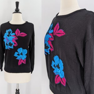 May include: Black crew neck sweater with a floral design. The sweater features large, stylized flowers in shades of blue and pink. The flowers are placed on the front of the sweater.