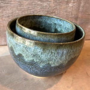 May include: Two nested ceramic bowls with a glossy, textured finish. The bowls have a gradient of colors, transitioning from a deep blue at the base to a teal and olive green at the rim. The bowls are handmade and suitable for serving or display.