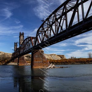 May include: A rusty metal bridge spans a river with a blue sky and white clouds in the background. The bridge has a series of vertical supports and a horizontal deck. The river is calm and reflects the sky.