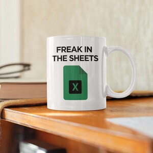 Könnte beinhalten: Weiße Keramiktasse mit dem Text "FREAK IN THE SHEETS" über einem grünen Excel-Symbol mit einem schwarzen "X". Die Tasse hat einen gebogenen Henkel und steht auf einer Holzoberfläche.