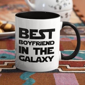 Op de afbeelding: Witte keramische mok met een zwarte binnenkant en handvat. De mok heeft de tekst "BEST BOYFRIEND IN THE GALAXY" in vette, zwarte letters. De mok staat op een patroon oppervlak.