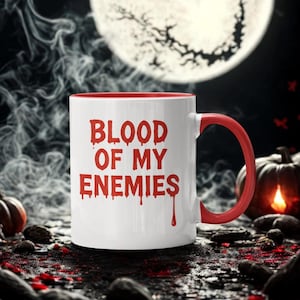 Op de afbeelding: Witte keramische mok met een rode binnenkant en handvat. De mok toont de tekst "BLOOD OF MY ENEMIES" in rode, druipende letters. De achtergrond omvat een volle maan, pompoenen en een donkere, spookachtige sfeer.