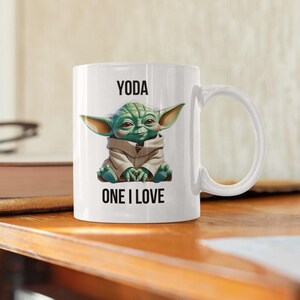 Op de afbeelding: Witte keramische mok met een groene Yoda-karakterillustratie en de tekst "YODA ONE I LOVE". De mok heeft een gebogen handvat en is geschikt voor warme dranken.