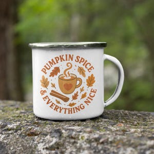 Op de afbeelding: Witte emaille mok met een zwarte rand. De mok heeft de tekst "Pumpkin Spice & Everything Nice" in oranje, samen met een koffiekopje, een kaneelstokje en herfstbladeren. Het heeft een wit handvat.