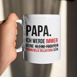Pode incluir: Caneca de cerâmica branca com o texto alemão "PAPA. ICH WERDE IMMER DEINE KLEINE TOCHTER FINANZIELLE BELASTUNG SEIN." em letras pretas e vermelhas. A caneca tem uma alça branca e é segurada por uma mão.