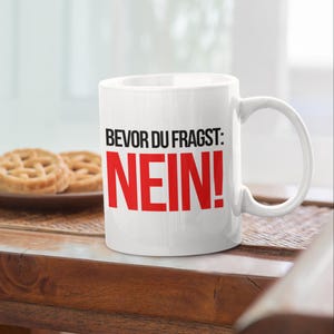 Könnte beinhalten: Weiße Keramik-Tasse mit dem Text "BEVOR DU FRAGST: NEIN!" in Schwarz und Rot. Die Tasse hat einen gebogenen Henkel und steht auf einer Holzoberfläche. Zwei kleine Gebäckstücke befinden sich auf einem Teller im Hintergrund.