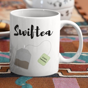 Op de afbeelding: Witte keramische mok met het woord "Swifttea" in zwart schrift. Een theezakje-afbeelding is op de zijkant gedrukt, met een groen label met de tekst "(Lisa's version)". De mok heeft een groot, gebogen handvat.