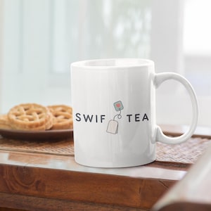Könnte beinhalten: Weiße Keramik-Tasse mit dem Text "SWIF TEA" und einer Teebeutel-Grafik. Die Tasse steht auf einem Holztisch mit einem Teller mit Keksen im Hintergrund. Die Tasse hat einen gebogenen Henkel.