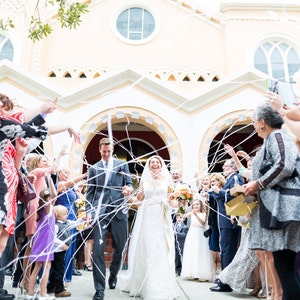 May include: A bride and groom are leaving their wedding ceremony, walking through a white ribbon tunnel. Guests throw white confetti as they exit the building. The bride is wearing a white lace dress and a white veil. The groom is wearing a dark suit and a white shirt. The ceremony is taking place in a white building with a large window.