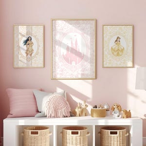 May include: Three framed art prints featuring Disney princess illustrations. The prints are in gold frames and hang on a pink wall. The artwork includes a princess, a castle, and another princess. Below the prints is a white bench with pillows and baskets.