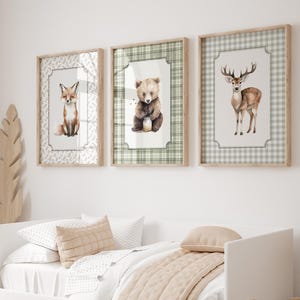 May include: Three framed watercolor prints of woodland animals: a fox, a bear, and a deer. The fox print has a white floral border, the bear print has a green plaid border, and the deer print has a gray plaid border. The frames are light brown.