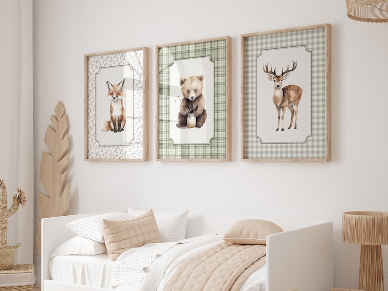 May include: Three framed watercolor prints of woodland animals: a fox, a bear, and a deer. The fox print has a white floral border, the bear print has a green plaid border, and the deer print has a gray plaid border. The frames are light brown.