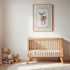 May include: A light wood baby crib with a mattress, a framed deer print, and teddy bears. The deer print features a watercolor illustration of a deer on a checkered background. The room has a neutral color palette.