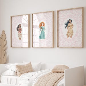 May include: Three framed art prints featuring illustrations of animated characters. Each print has a light wood frame and a pink damask background. The characters are in oval frames. The prints are hung above a white bed.
