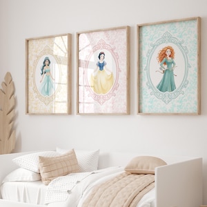May include: Three framed art prints featuring watercolor illustrations of princesses. Each princess is depicted within an oval frame against a patterned background. The frames are displayed above a white bed with neutral-toned bedding.