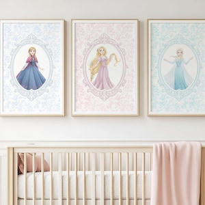 May include: Three framed art prints featuring illustrations of princesses. Each print has a different princess in an oval frame, set against a patterned background. The frames are gold-colored, and the prints are displayed above a crib.