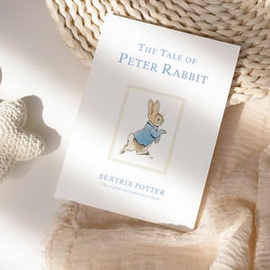 Puede incluir: Un ejemplar de "The Tale of Peter Rabbit" de Beatrix Potter. La portada del libro presenta el t&iacute;tulo en azul claro, una ilustraci&oacute;n de Peter Rabbit y el nombre del autor. Tambi&eacute;n se ven un juguete blanco en forma de estrella y una cesta de mimbre.