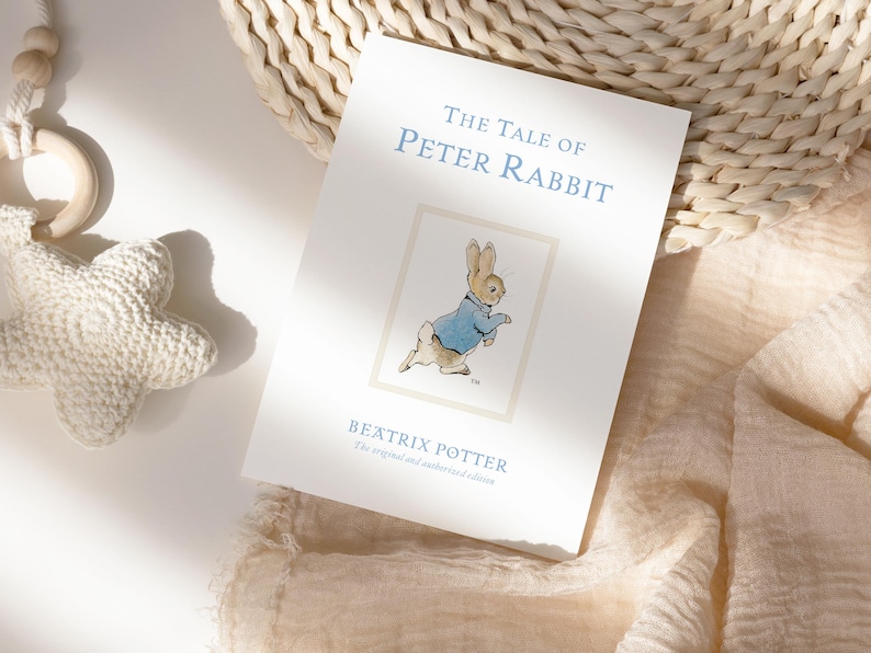 Puede incluir: Un ejemplar de "The Tale of Peter Rabbit" de Beatrix Potter. La portada del libro presenta el t&iacute;tulo en azul claro, una ilustraci&oacute;n de Peter Rabbit y el nombre del autor. Tambi&eacute;n se ven un juguete blanco en forma de estrella y una cesta de mimbre.