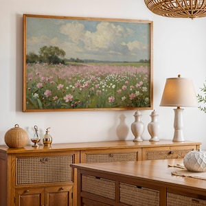 Peut inclure: Une peinture encadr&eacute;e repr&eacute;sente un paysage avec un champ de fleurs roses et blanches sous un ciel nuageux. Le tableau est accroch&eacute; au-dessus d'un buffet en bois avec des panneaux tress&eacute;s. Une lampe avec un abat-jour beige et des vases d&eacute;coratifs sont sur le buffet.
