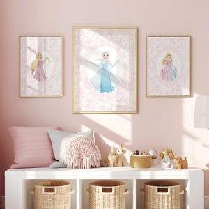 May include: Three framed princess art prints with gold frames hang on a pink wall. The prints feature illustrations of princesses in oval frames against a pink patterned background. Below, a white shelf holds pillows, baskets, and decorative items.