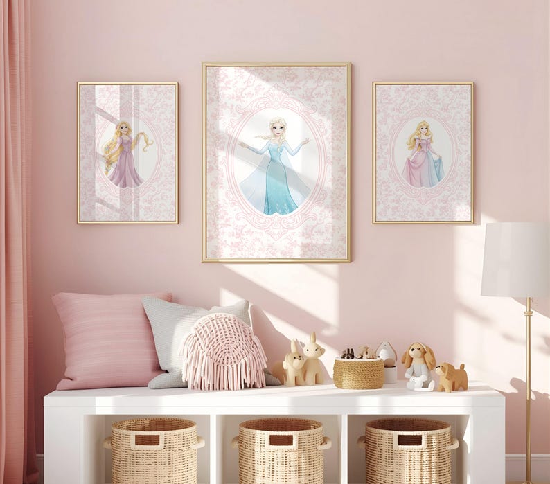 May include: Three framed princess art prints with gold frames hang on a pink wall. The prints feature illustrations of princesses in oval frames against a pink patterned background. Below, a white shelf holds pillows, baskets, and decorative items.