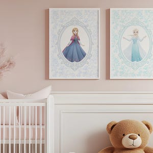 May include: Two framed prints featuring Disney princesses, Anna and Elsa, in ornate oval frames. The prints are set against a light blue floral patterned background. The room has a white crib, a brown teddy bear, and pink accents.