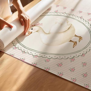 Peut inclure: Un rouleau de papier peint en cours de d&eacute;roulement, r&eacute;v&eacute;lant un motif fantaisiste. Le papier peint pr&eacute;sente une grande illustration d'une oie blanche avec un n&oelig;ud rose, dans un cadre ovale. Le fond est &agrave; rayures roses et &agrave; petits motifs de roses.