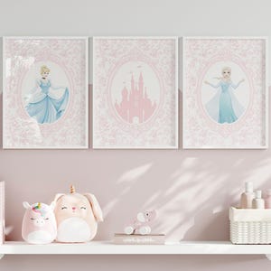 May include: Three framed art prints featuring Cinderella, a pink castle, and Elsa. Each print is set against a pink floral background within a white frame. The prints are displayed above a shelf with plush toys and decorative items.