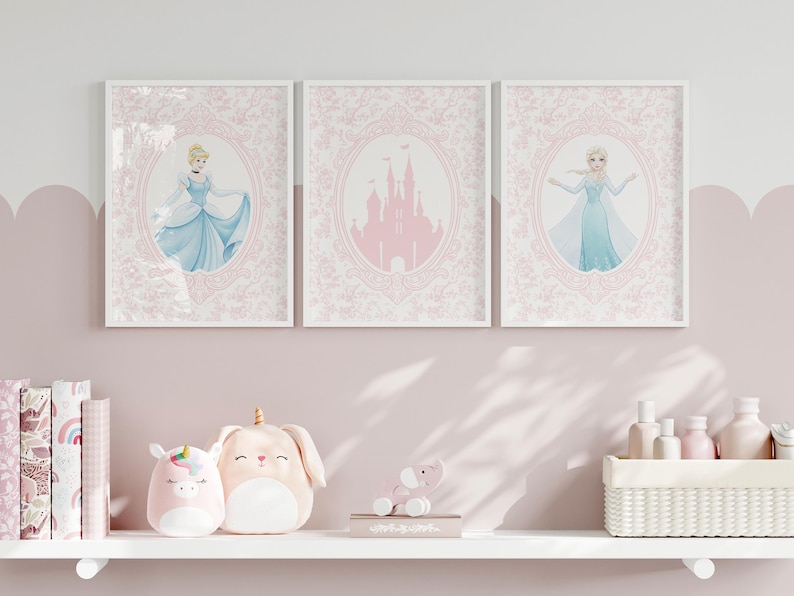 May include: Three framed art prints featuring Cinderella, a pink castle, and Elsa. Each print is set against a pink floral background within a white frame. The prints are displayed above a shelf with plush toys and decorative items.