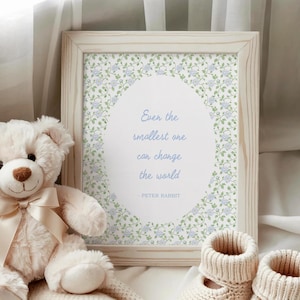 Puede incluir: Un cuadro enmarcado con la cita "Incluso el m&aacute;s peque&ntilde;o puede cambiar el mundo" de Peter Rabbit. Un oso de peluche blanco con una cinta crema y un par de botines de beb&eacute; est&aacute;n junto al marco. La impresi&oacute;n tiene un dise&ntilde;o floral.
