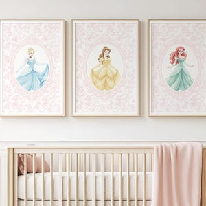 May include: Three framed art prints featuring watercolor illustrations of princesses in elegant gowns. Each princess is set against a pink floral patterned background within an oval frame. The prints are displayed above a wooden crib with a pink blanket.