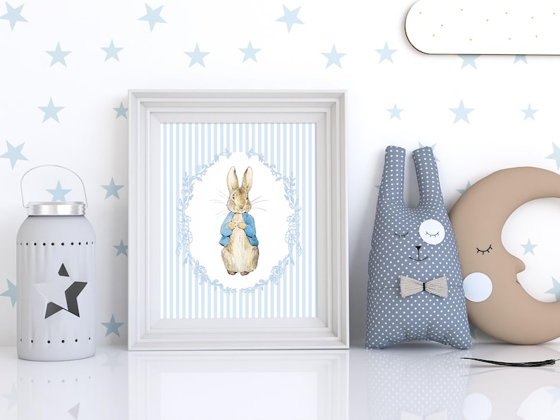 Puede incluir: Una impresi&oacute;n enmarcada con una ilustraci&oacute;n en acuarela de un conejo con una chaqueta azul, sobre un fondo a rayas azules y blancas. Junto a ella, un peluche de conejo gris con pajarita, una luna creciente y una linterna en forma de estrella.