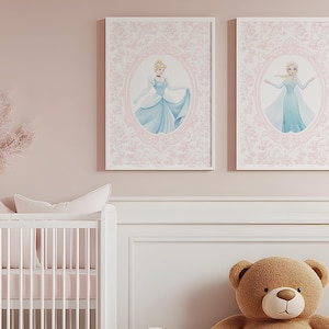 May include: Two framed prints featuring illustrations of princesses in blue gowns, set against a pink floral background. The prints are displayed above a white crib and a large teddy bear, creating a soft, feminine nursery theme.