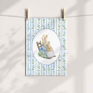 Puede incluir: Una impresi&oacute;n en acuarela que representa dos conejos, uno adulto y otro ni&ntilde;o, sentados en una peque&ntilde;a silla. La impresi&oacute;n est&aacute; enmarcada en un &oacute;valo sobre un fondo de rayas verticales azules y blancas con detalles florales. La impresi&oacute;n est&aacute; sujeta con pinzas de madera.