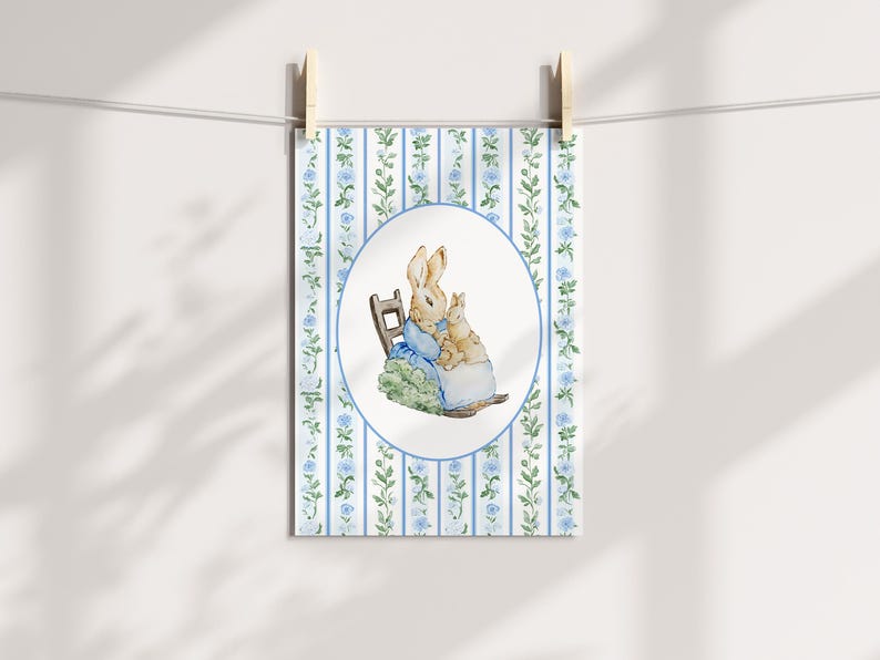 Puede incluir: Una impresi&oacute;n en acuarela que representa dos conejos, uno adulto y otro ni&ntilde;o, sentados en una peque&ntilde;a silla. La impresi&oacute;n est&aacute; enmarcada en un &oacute;valo sobre un fondo de rayas verticales azules y blancas con detalles florales. La impresi&oacute;n est&aacute; sujeta con pinzas de madera.