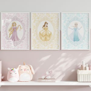 May include: Three framed princess art prints. Each features a different princess in an oval frame with a floral background. The prints are in pastel colors and are displayed above a shelf with toys and books.