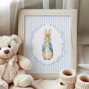 Puede incluir: Una impresi&oacute;n enmarcada con una ilustraci&oacute;n en acuarela de Peter Rabbit con una chaqueta azul, sobre un fondo a rayas azules y blancas. Un oso de peluche blanco con una cinta crema y botines de beb&eacute; de punto est&aacute;n en primer plano.