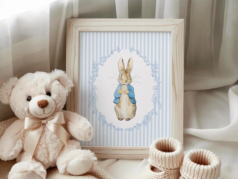 Puede incluir: Una impresi&oacute;n enmarcada con una ilustraci&oacute;n en acuarela de Peter Rabbit con una chaqueta azul, sobre un fondo a rayas azules y blancas. Un oso de peluche blanco con una cinta crema y botines de beb&eacute; de punto est&aacute;n en primer plano.